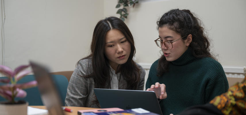 Two academics sitting on a sofa, looking at a laptop. The person on the right is speaking and gesturing at the screen.