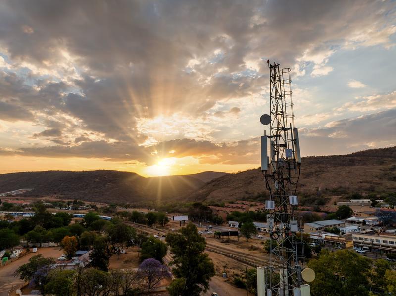 An aerial view of a cellular antenna at sunset, in front of a town, railway line and some hills.