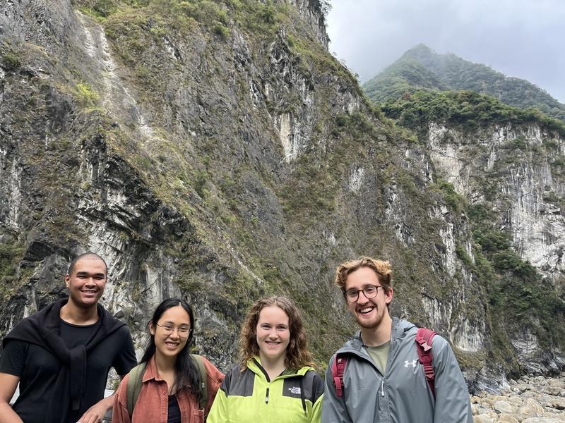 Four students, dressed in outdoor clothing and carrying rucksacks, standing in front of a rocky cliff face with a mountain behind them.