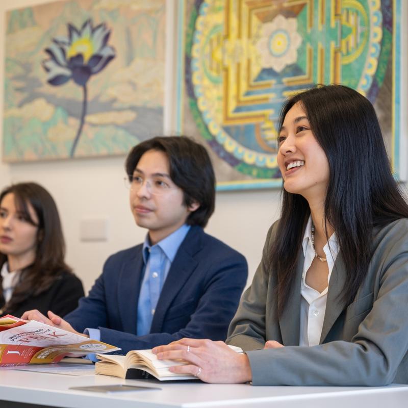 Three students sitting in a row behind a table in a seminar. There are three colourful abstract paintings behind them. 