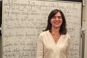 Lia Chokoshvili has shoulder-length dark hair. She is wearing a white v-neck blouse and standing in front of a whiteboard covered in Georgian script.