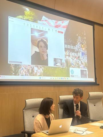 Lia Chokoshvili sits at a large wooden desk, looking at a man to her side who is speaking into a microphone. Behind them is a large screen, where Tamta Melashvili is appearing via video-call.