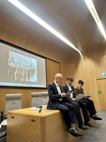 Three men sitting on the edge of a large wooden desk. They are wearing smart suits, holding large books and reading aloud. Behind them is a screen showing a black and white photo of a group of women.
