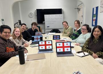 A group of eight students sat around a desk, facing and smiling at the camera. In front of them are laptops showing the Georgian flags