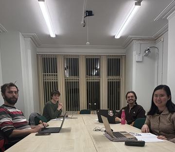 A group of four students sat a a long desk, with laptops and papers. They are facing the camera and smiling