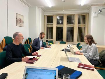Three students sitting around a long table, looking at laptops and papers while smiling. 