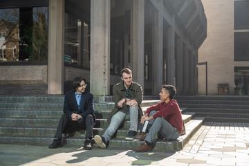 Three students sitting on steps outside a building with large grey pillars.