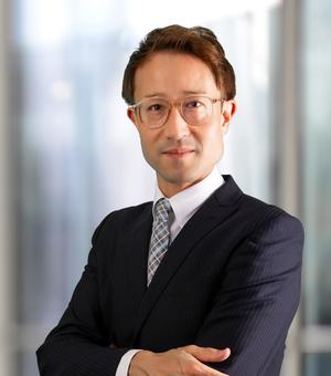 Dr Buchmeier has short brown hair and is wearing clear-rimmed glasses. He is wearing a pinstripe suit, shirt and tie. He is standing with his arms folded, looking at the camera. 