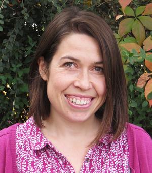 Jo Pready has shoulder length brown hair. She is wearing a pink patterned blouse and pink cardigan and smiling. 