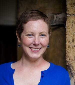 Dr LaPorte is leaning against a wall and smiling. She has cropped brown hair and is wearing a bright blue cardigan and blue earrings. 