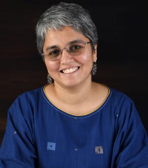 Janaki Srinivasan has short grey hair and glasses. She is wearing a navy blue dress with embroidered details around the neckline, and large silver earrings.