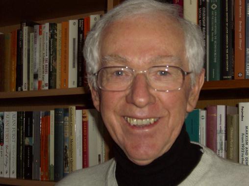 Alan has grey hair, glasses and is smiling at the camera. He's wearing a black shirt under a cream coloured jumper. He's standing in front of a bookcase.