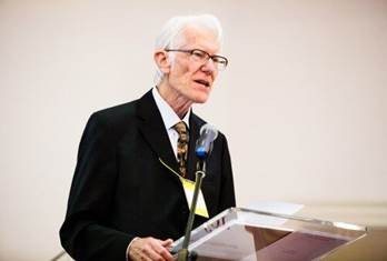 Arthur Stockwin has grey hair and wearing glasses. He's standing at a podium speaking into a microphone. He's wearing a white shirt, tie and smart black jacket. 