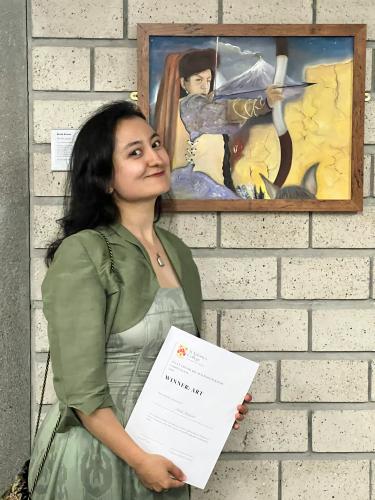 akida anwar has long dark hair and wearing a pale green dress with cropped jacket. She is holding a certificate and standing next to her framed painting. The painting shows an archer on horseback and aiming their bow and arrow. There is a smoking volcano behind the archer on horseback.