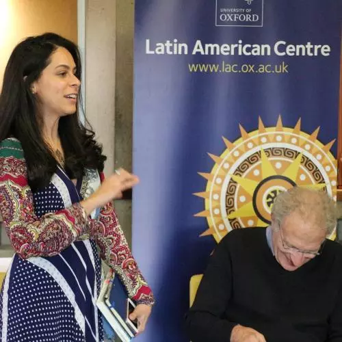 A woman with long dark hair wearing a colourful patterned dress presents in front of an event banner. 2022