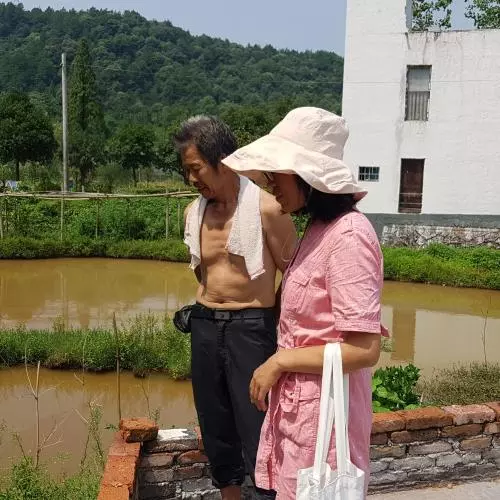 A women and a man looking into a large pond filled with brown water. The pond by surrounded by grass, and a concrete pathway. 