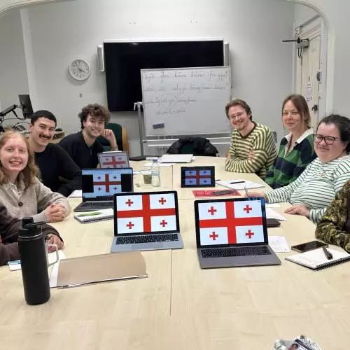 A group of eight students sat around a desk, facing and smiling at the camera. In front of them are laptops showing the Georgian flags