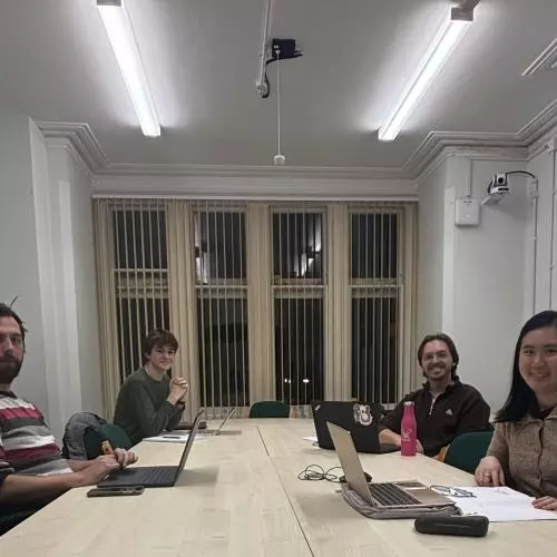 A group of four students sat a a long desk, with laptops and papers. They are facing the camera and smiling