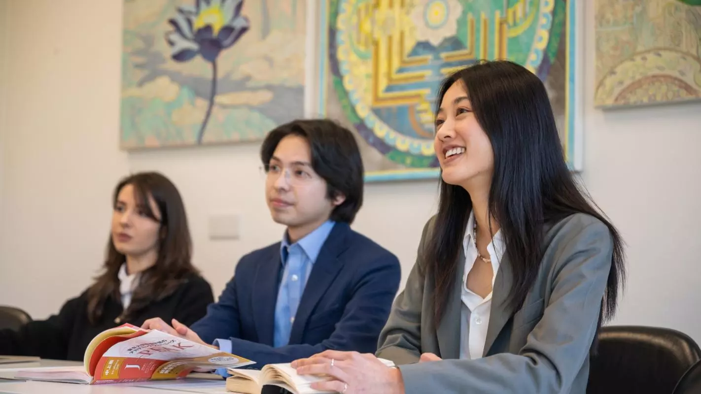 Three students sitting in a row behind a table in a seminar. There are three colourful abstract paintings behind them. Photo credit: John Cairns Photography