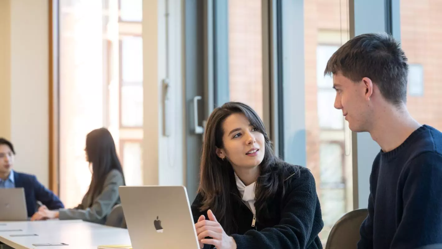 Two students sitting at a desk, looking at a laptop between them. The female student, sat on the left, is looking to the other student and explaining something. Photo credit: John Cairns Photography