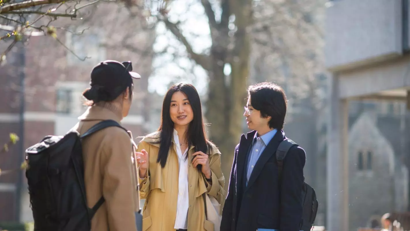 Three students standing outside chatting. They are wearing light jackets and carrying backpacks and books. Photo credit: John Cairns Photography