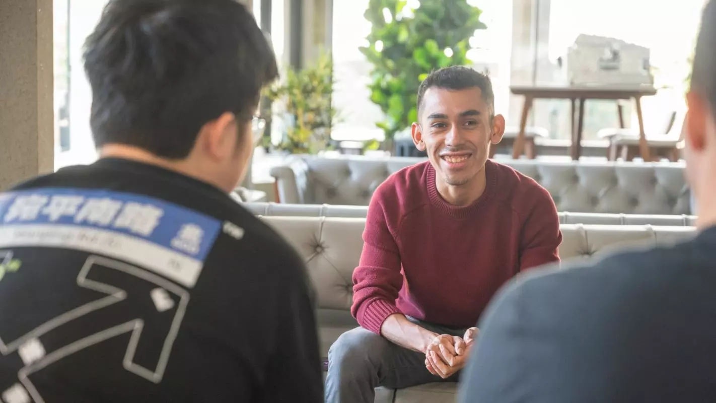 A male student wearing a red jumper sitting on a sofa in a shared lounge area. He is talking to two other students with their backs to the camera. 