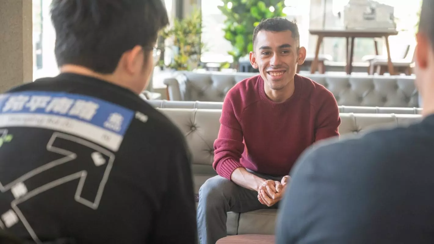 A male student wearing a red jumper sitting on a sofa in a shared lounge area. He is talking to two other students with their backs to the camera. 