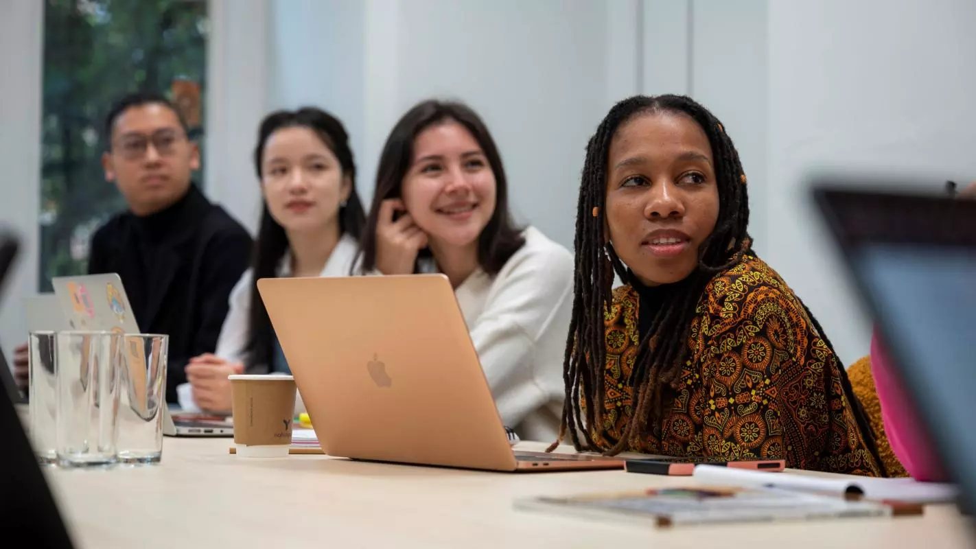 A group of students in a seminar, sat in a line at a desk with laptops. 