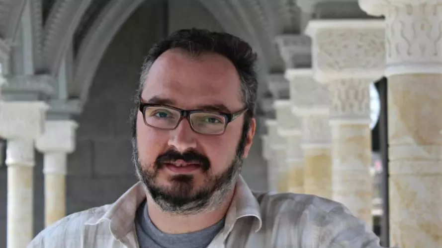 David has short dark hair, short beard and glasses. He is standing beneath a row of ornate stone arches