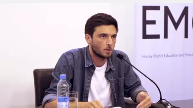 Dr Eristavi has short dark hair and is wearing a denim shirt. He is sat at a desk and speaking into a microphone