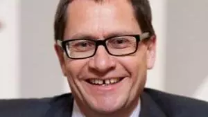 Roger Goodma has short dark hair and dark glasses. He is wearing a shirt, tie and dark blazer