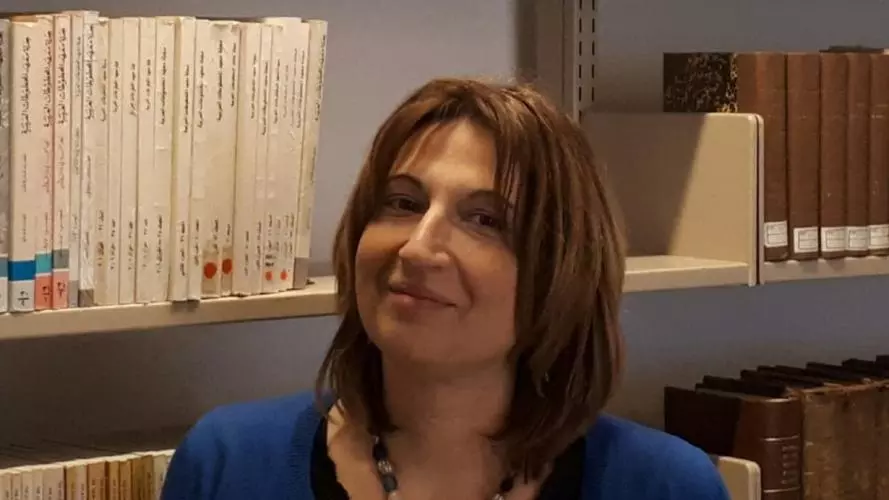 Tamara Pataridze has shoulder-length brown hair and is wearing a dark dress with a blue cardigan. She is holding a book open and standing in front of bookshelves