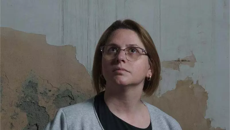 Tea Kamushadze has short brown hair and glasses. She is wearing a grey cardigan and black dress and standing in front of a distressed wall