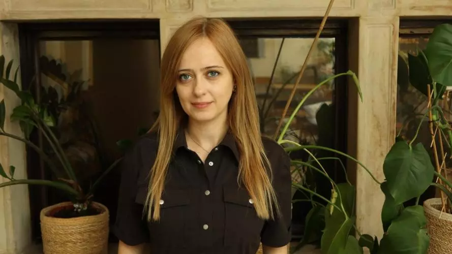 Esra Akgemci has long blonde hair. She is wearing a black jumpsuit and standing in front of some plants