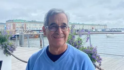 Jesus Velasco has short grey hair and glasses. He is wearing a light blue jumper and sat at a waterfront