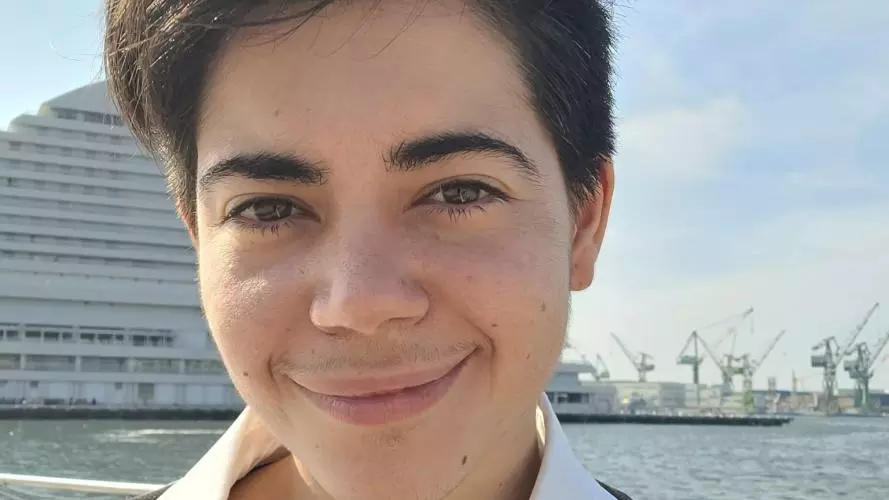 Pan has short dark hair. They are wearing a black and white shirt with a large collar, standing in front of a harbour