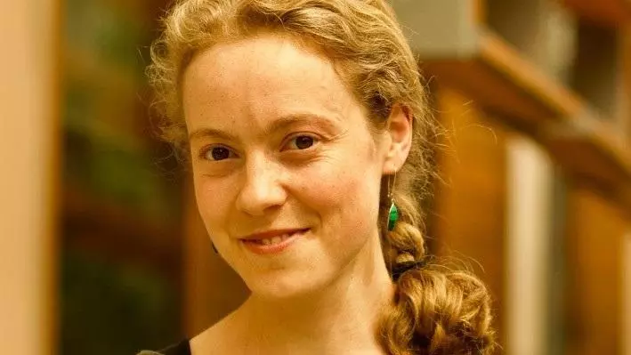 Dr Driessen has curly fair hair tied in a plait. She is wearing a green sweater
