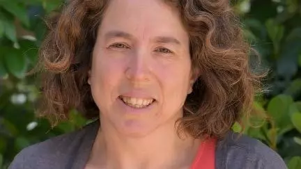 Tali has curly brown shoulder-length hair and she is smiling at the camera. She's wearing a salmon pink t shirt under a grey cardigan.  