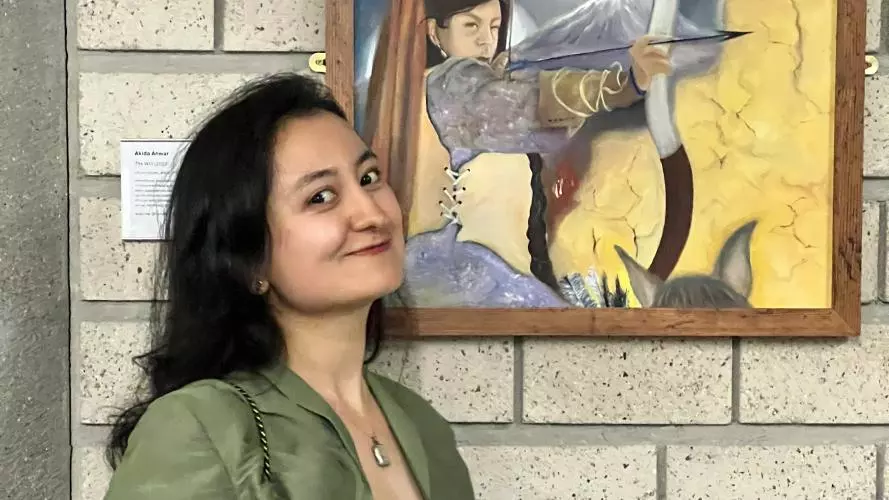 akida anwar has long dark hair and wearing a pale green dress with cropped jacket. She is holding a certificate and standing next to her framed painting. The painting shows an archer on horseback and aiming their bow and arrow. There is a smoking volcano behind the archer on horseback.