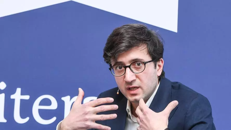 Professor David Maziashvili has dark brown hair, wearing a white shirt under a navy blue jacket. He's talking and gesturing with his hands. He's sitting down with a 'London Book Fair' backdrop behind him.