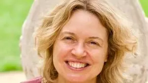 Rachel has shoulder-length blonde wavy hair and is smiling at the camera. She's wearing a ruby coloured top with silver pendant necklace
