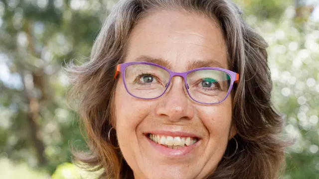 Rita Abrahamsen has brown shoulder-length hair and wearing purple glasses. She's wearing a navy blue top and smiling at the camera 