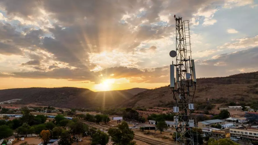 An aerial view of a cellular antenna at sunset, in front of a town, railway line and some hills.
