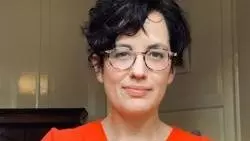 Maria has short dark hair and glasses, and is wearing a bright orange v-neck top