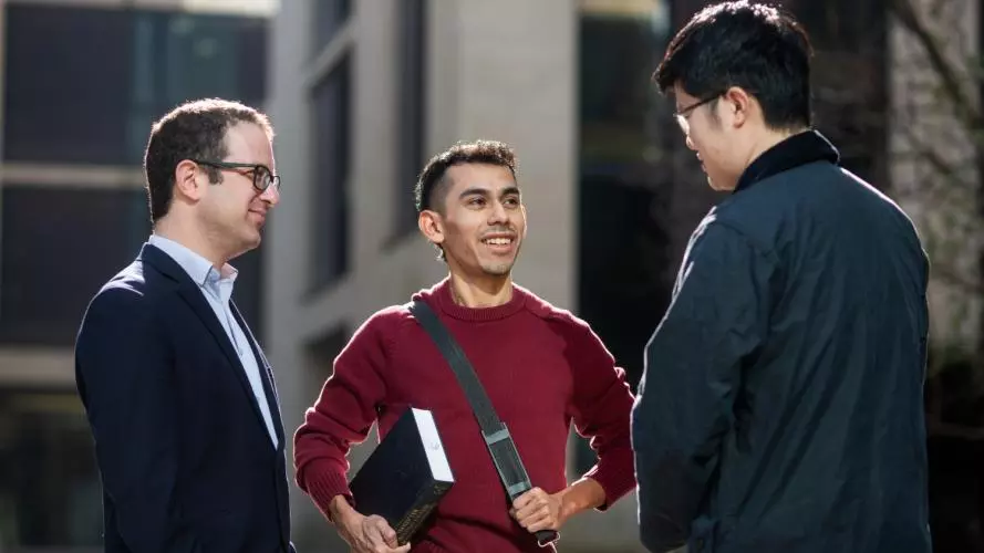 Two students and a teacher in a group, standing and chatting outside department buildings. 
