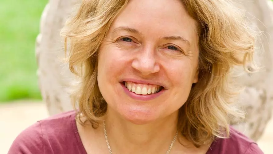 Rachel has shoulder-length blonde wavy hair and is smiling at the camera. She's wearing a ruby coloured top with silver pendant necklace