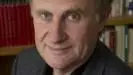 William has short brown hair and is wearing a black shirt with dark grey jacket. There is a bookcase behind him. 