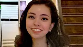 Eliza has shoulder length brown hair and brown eyes. She is wearing a dark blue silk shirt, brown checked blazer and gold jewellery. She is smiling at the camera. 