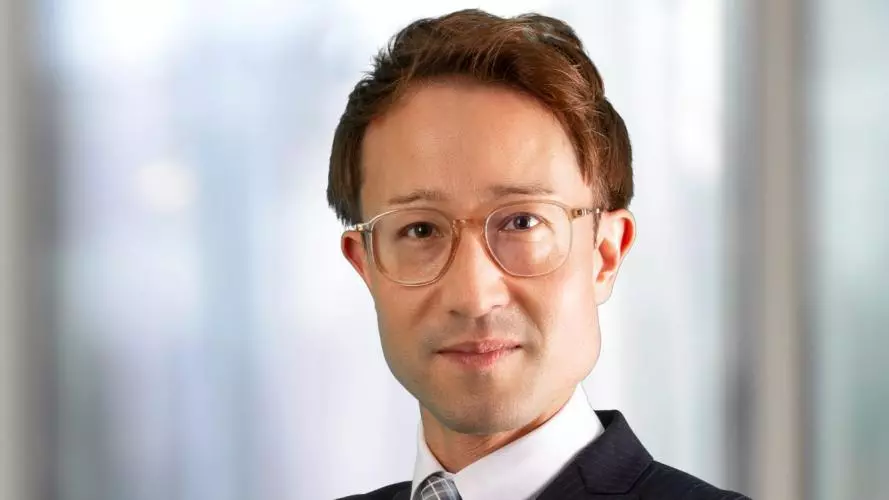 Dr Buchmeier has short brown hair and is wearing clear-rimmed glasses. He is wearing a pinstripe suit, shirt and tie. He is standing with his arms folded, looking at the camera. 