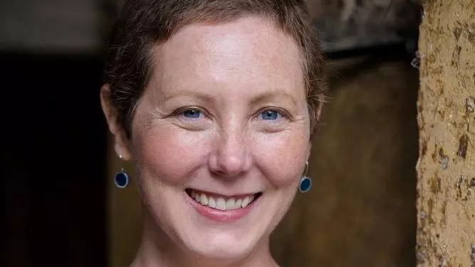 Dr LaPorte is leaning against a wall and smiling. She has cropped brown hair and is wearing a bright blue cardigan and blue earrings. 